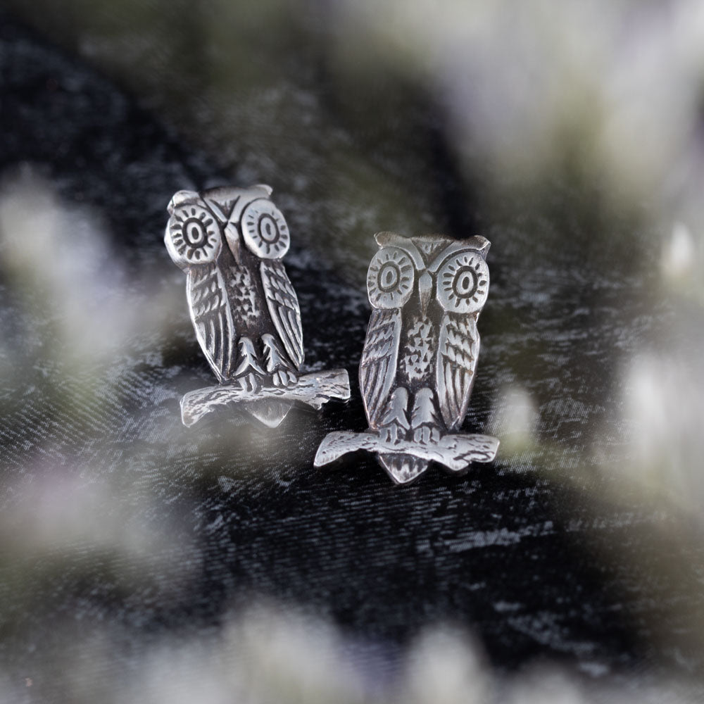 Pair of sterling silver owl earrings with intricate etched details, perched on branches, displayed on dark textured cloth with blurred lavender sprigs in the foreground.