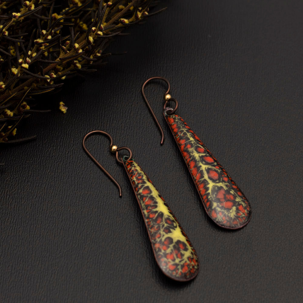 Long teardrop-shaped enamel earrings in fiery shades of red, yellow, and black with organic patterns, displayed on a dark gray background with autumn sprigs.