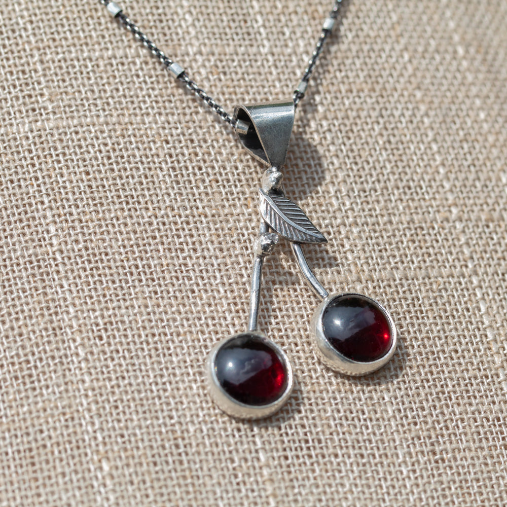 Close view of a handmade garnet cherry necklace with sterling silver stems and a detailed silver leaf. Photo taken outside,