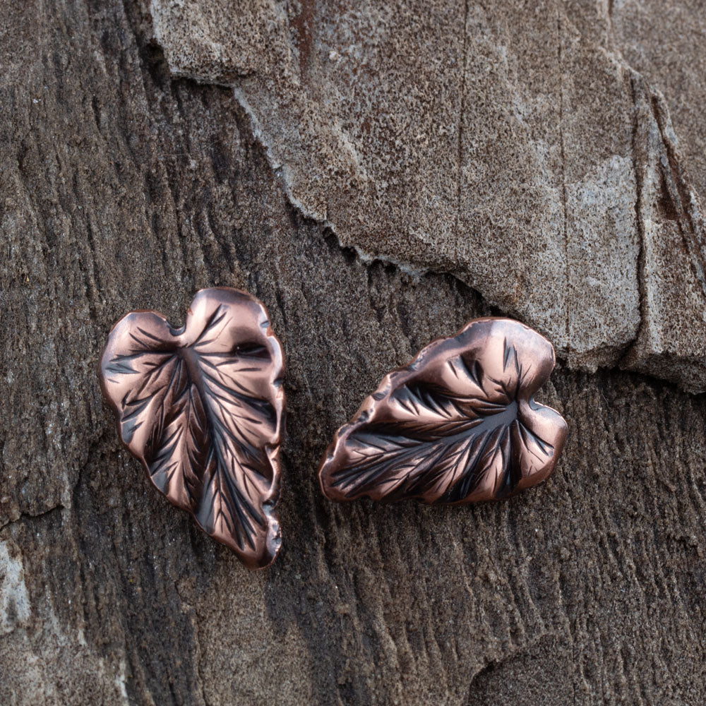 Handcrafted copper leaf post earrings with oxidized details, featuring realistic vein texture and asymmetrical heart-shaped foliage design on rustic wood background.