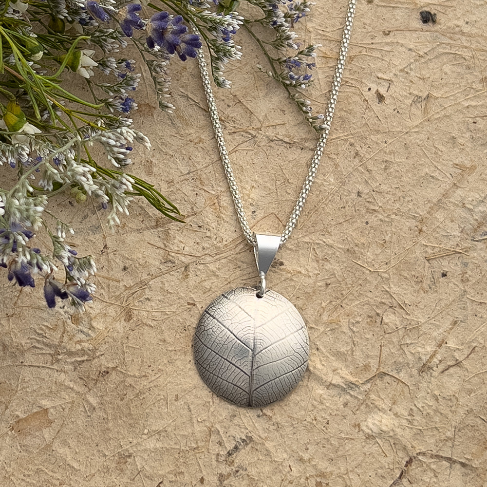 Round silver pendant with a delicate leaf vein imprint, hanging on a textured chain, displayed on a neutral natural background with dried lavender and white wildflowers.