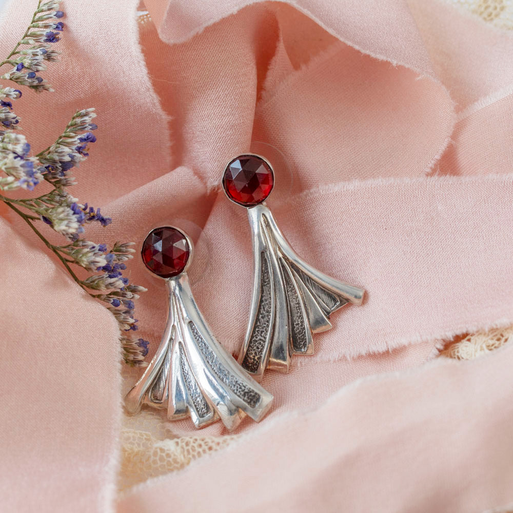 Art Deco-inspired sterling silver earrings featuring deep red faceted garnet stones and flared geometric fan shapes, styled with pink silk and lavender floral elements for a feminine luxe vibe.