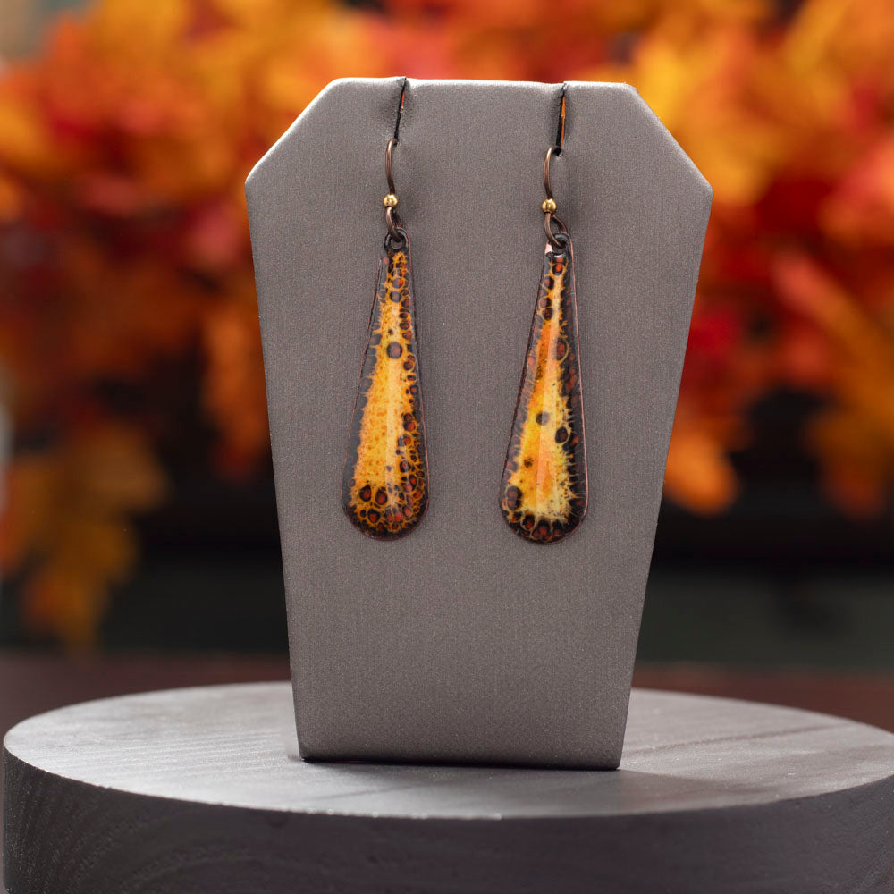 Long teardrop enamel earrings in warm tones of orange, yellow, and black with a speckled pattern, displayed on a display with blurred autumn leaf background.