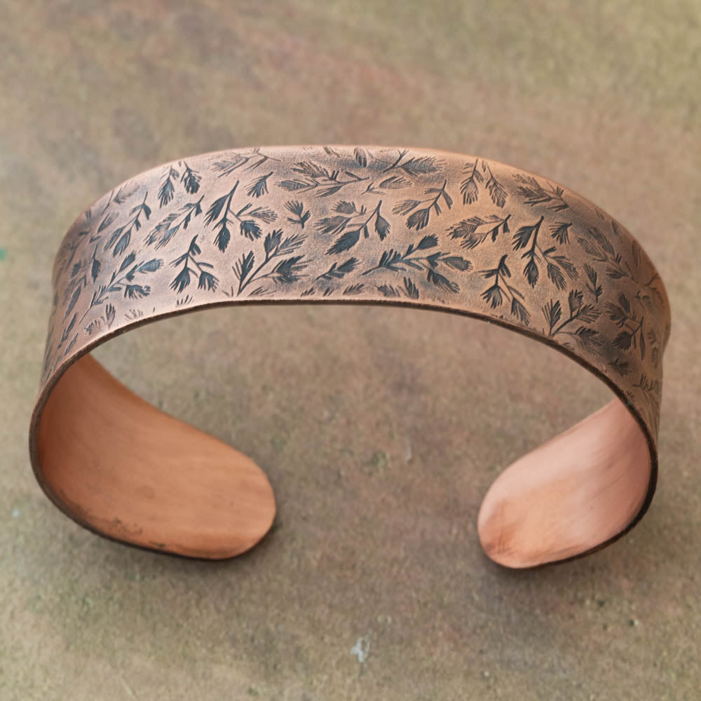Anticlastic copper cuff bracelet with evergreen sprig pattern on a textured background.