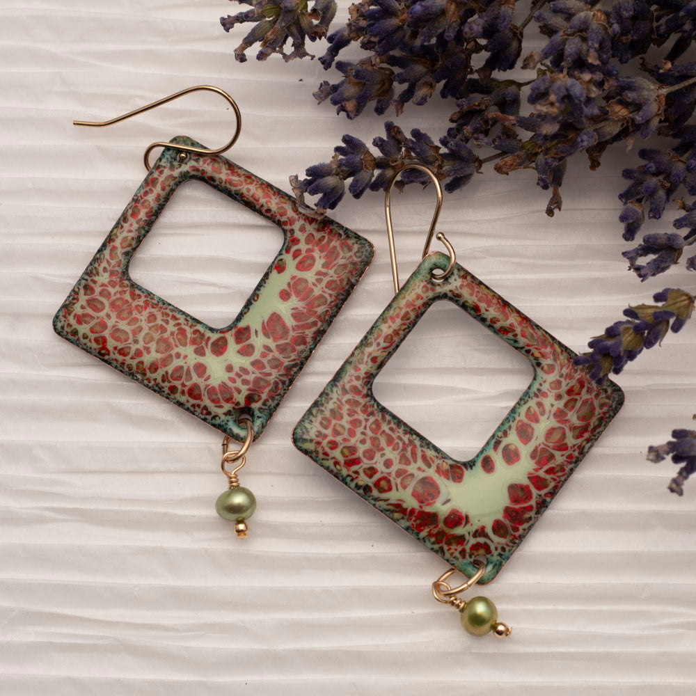 Square copper enamel earrings in mint green with red and black crackle design, finished with green pearl dangles and gold ear wires, styled on white textured paper with sprigs of lilac.