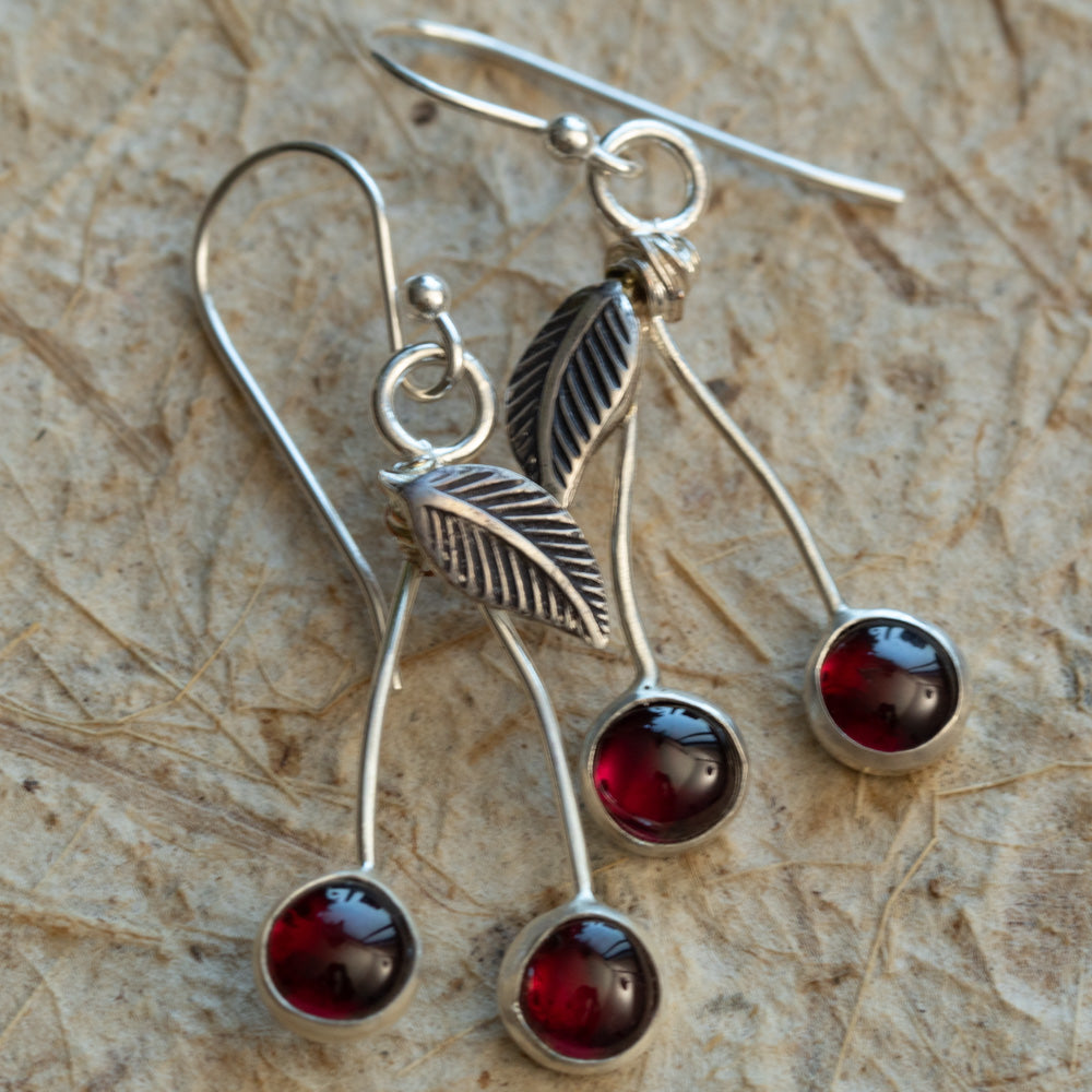Garnet cherry earrings handmade in sterling silver with curved stems and textured leaves on a warm neutral background.