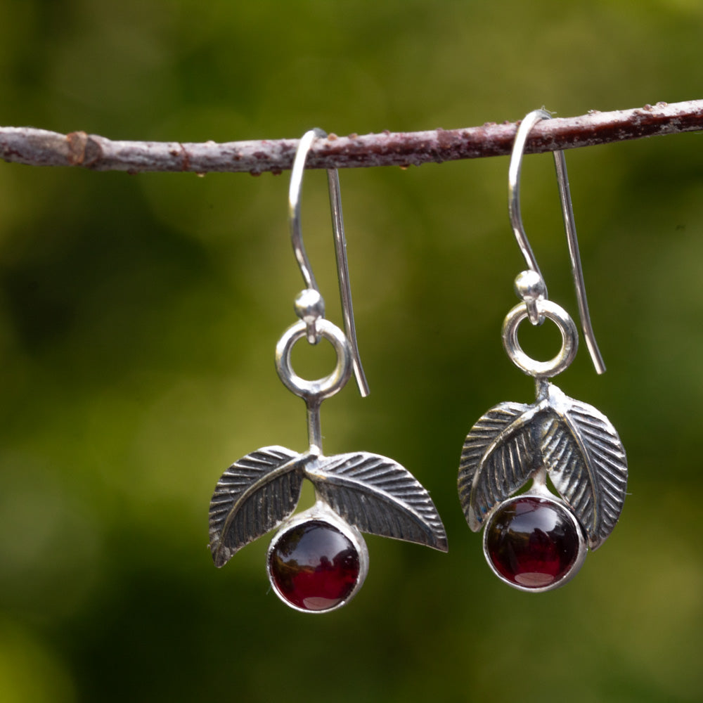 Close-up of garnet cherry earrings handmade in sterling silver with oxidized textured leaves hanging from a branch.