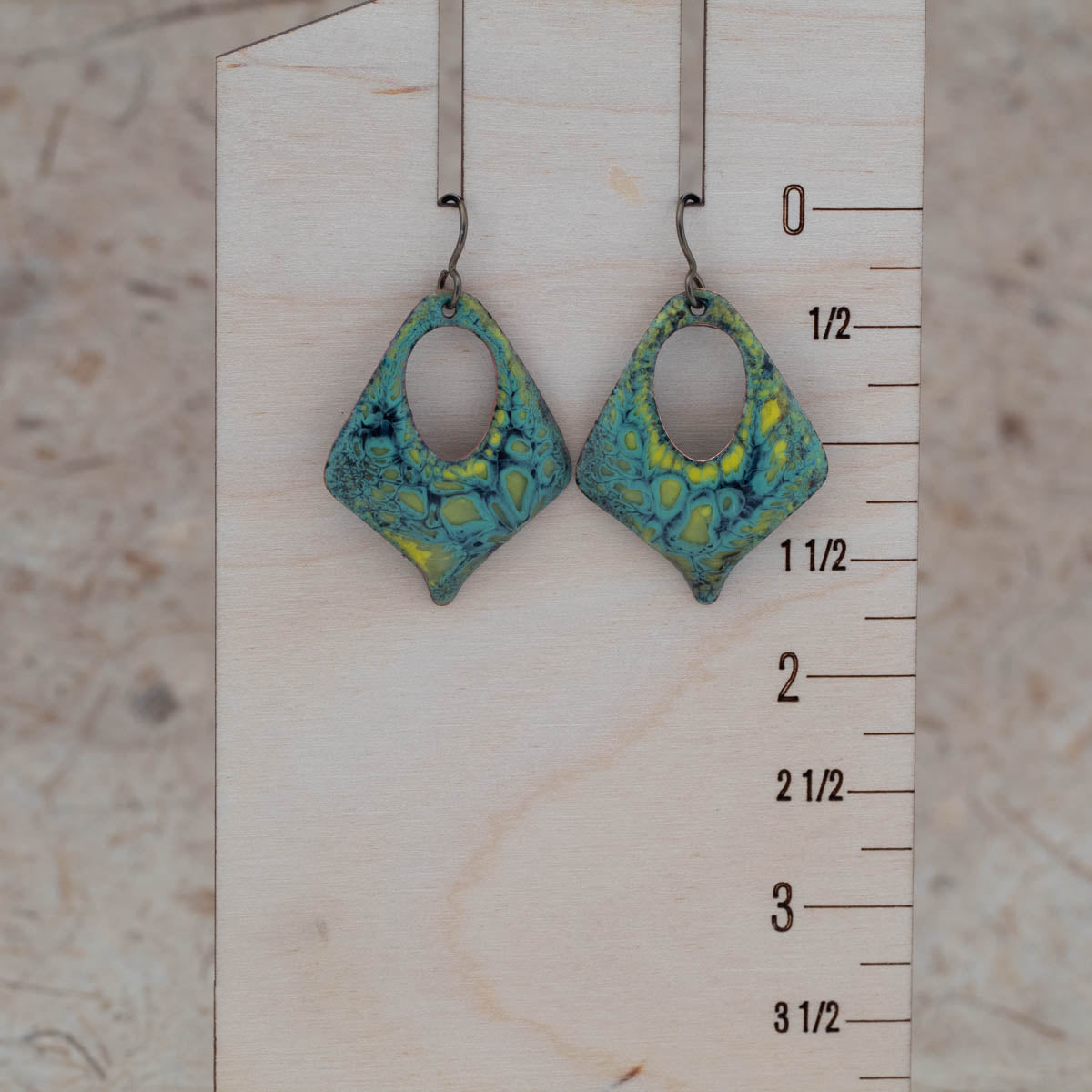 Handmade diamond-shaped enamel earrings with earthy green, yellow, and teal tones in a swirling pattern. Open oval center design with rustic patina, hanging on dark metal ear wires. Displayed on birch bark with wildflowers – Moonlight Earrings by Divella Designs.