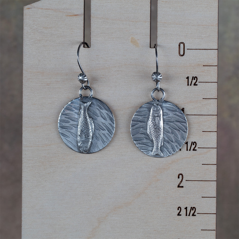 Handcrafted sterling silver earrings featuring detailed fish motifs swimming over textured water-like backgrounds – Swimmers artisan earrings by Divella Designs, displayed on a wooden ruler for scale