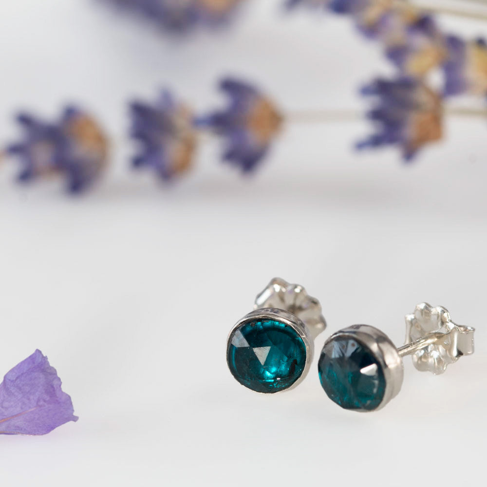 Close up of sterling silver stud earrings featuring faceted teal kyanite gemstones, photographed on a white background with soft lavender accents.