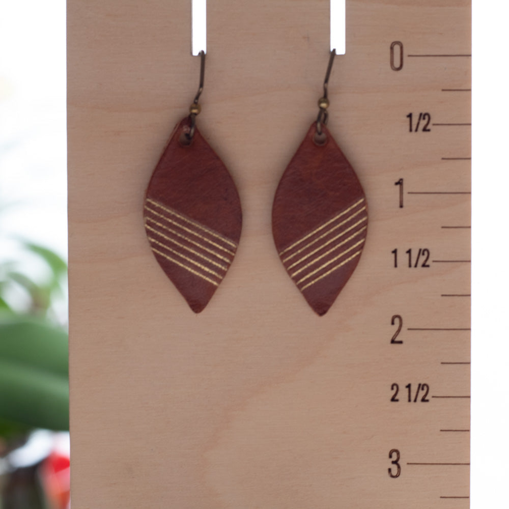 A pair of teardrop-shaped wooden dangle earrings with gold-toned horizontal stripes, hanging from a wooden display block with a size ruler visible on the right.