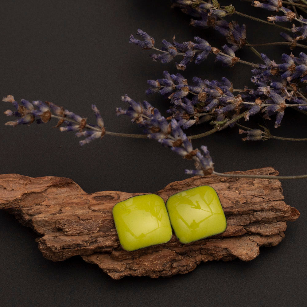 A pair of green enamel stud earrings on a wooden surface with a backdrop of purple flowers.