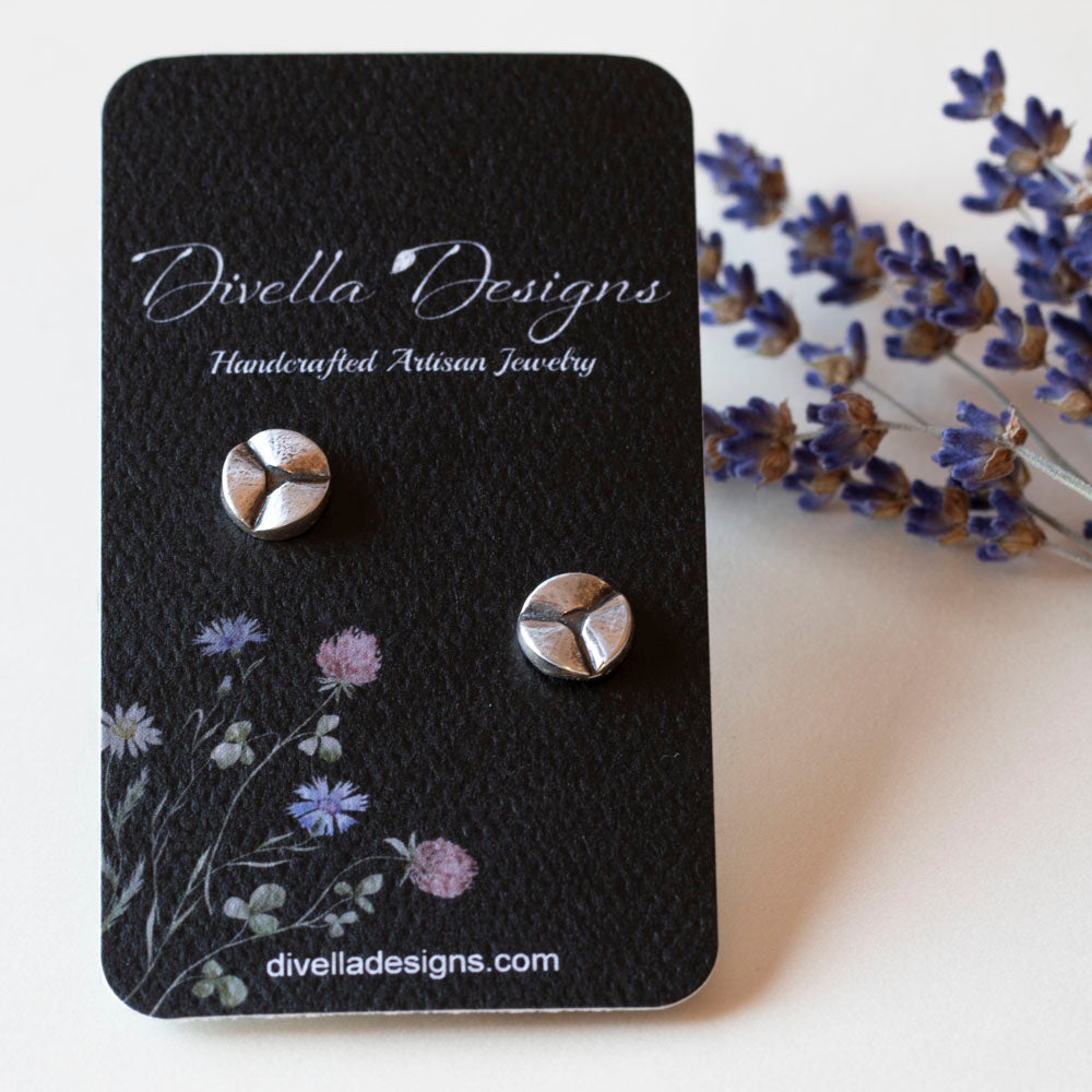 Modern minimalist sterling silver stud earrings with a three-pronged abstract design, displayed on a black branded card on a background with sprigs of dried lavender.