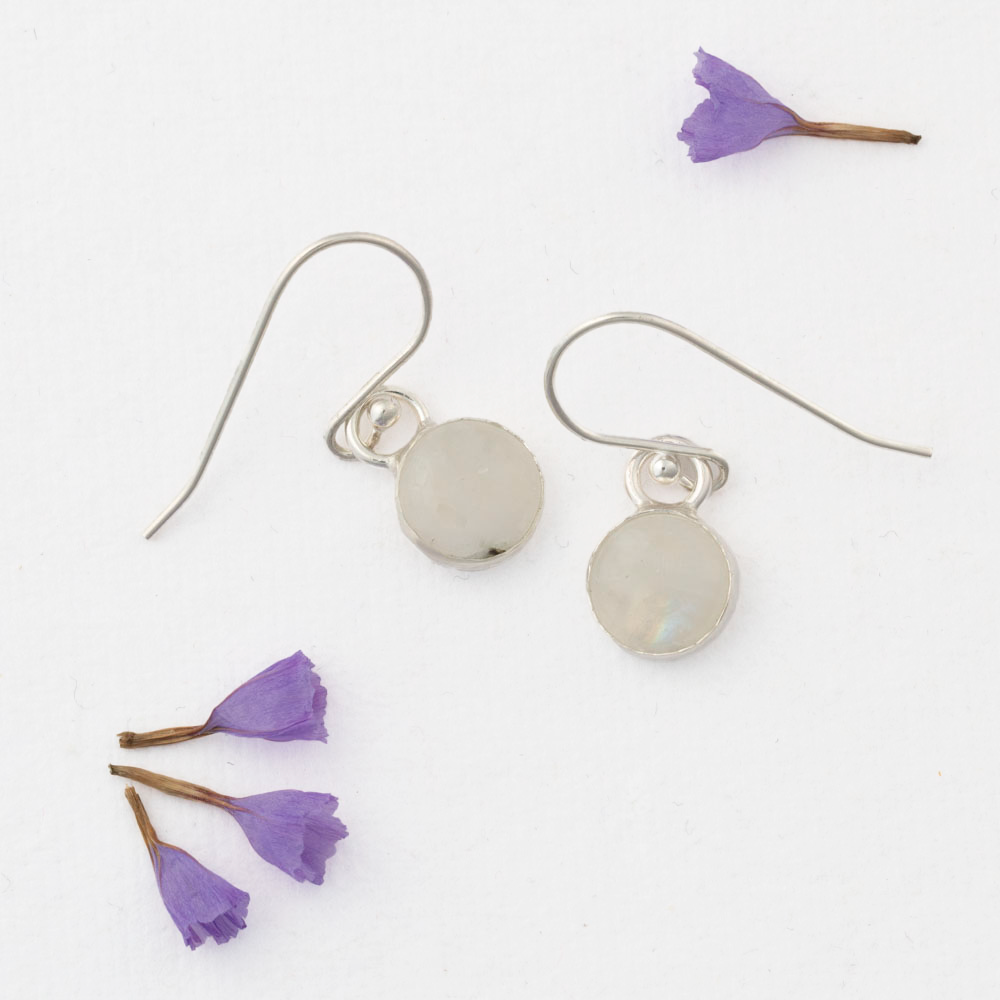 Delicate moonstone drop earrings in sterling silver with round cabochon stones, simple handmade dangles displayed with scattered purple flower petals on white background