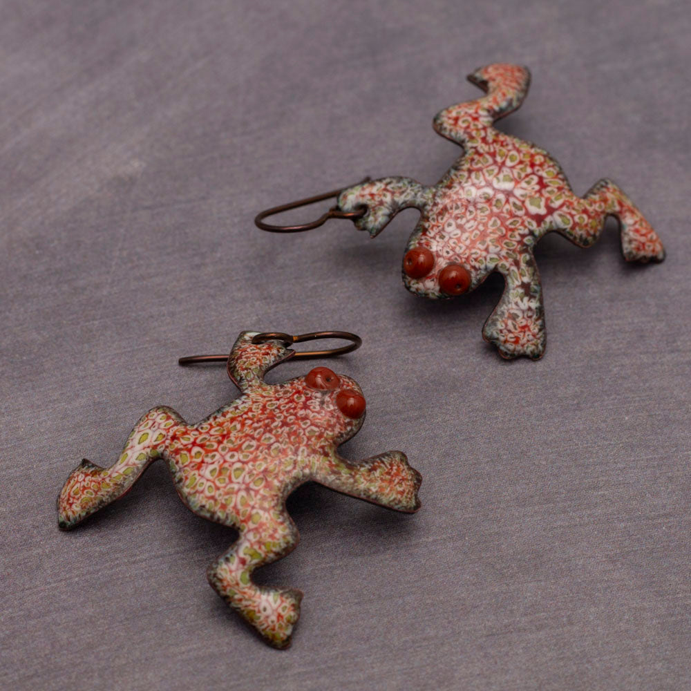 Playful red and green enamel frog-shaped earrings with dotted patterns and raised spiral eyes, displayed on a gray stand against a light gray background.