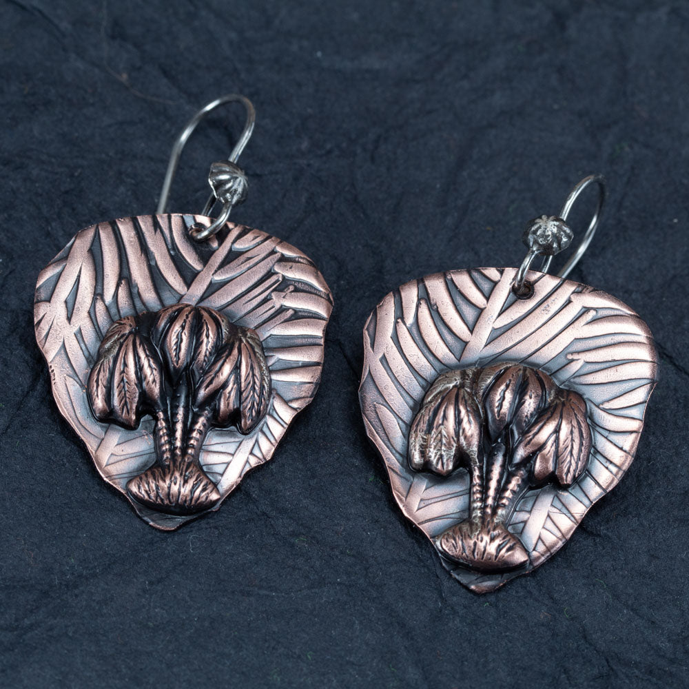 Handcrafted copper earrings with sculpted palm trees layered over embossed leaf-patterned backdrops, tropical artisan jewelry by Divella Designs – displayed on dark slate background.