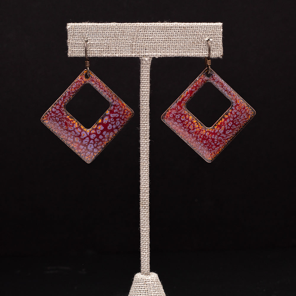 Square enamel earrings in rich plum and orange tones with speckled detail, styled on an earrings display against a black background.