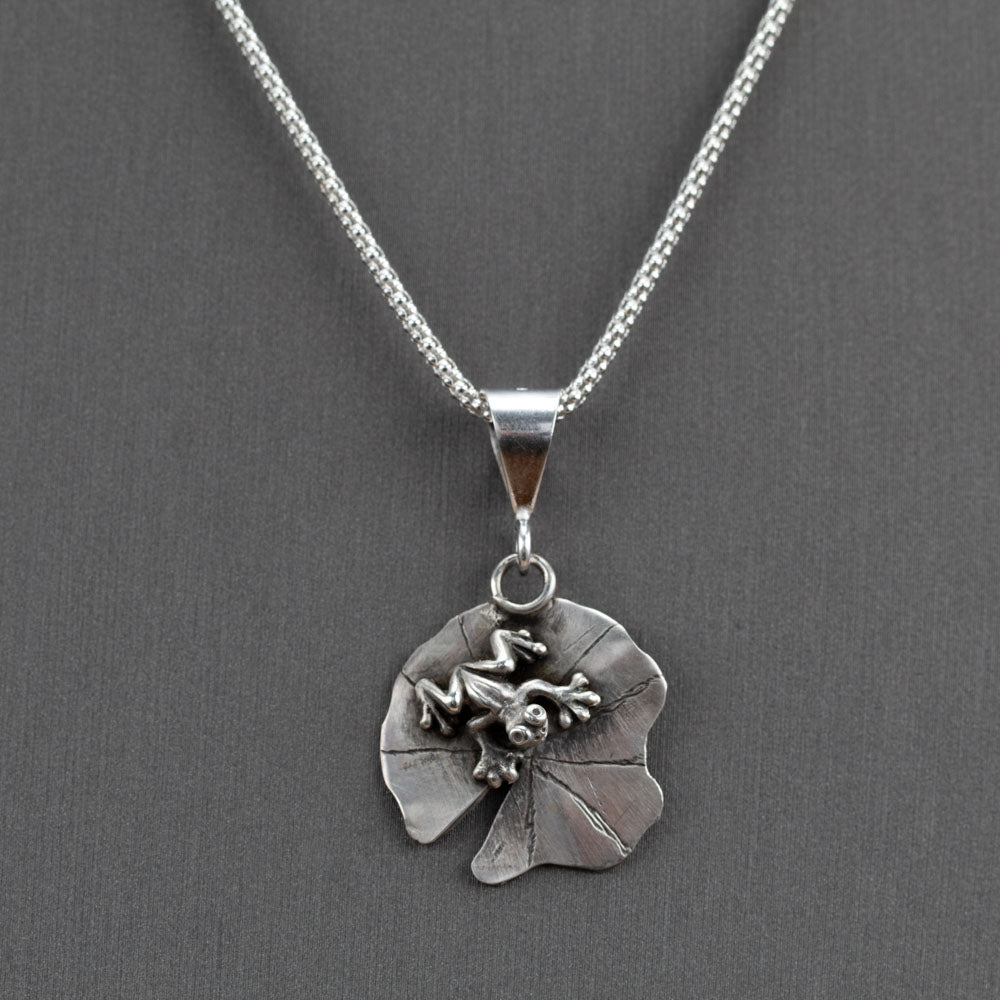 Handcrafted sterling silver frog pendant resting on a textured lily pad, nature-inspired Pond Jumper necklace by Divella Designs – shown on gray background with popcorn chain.