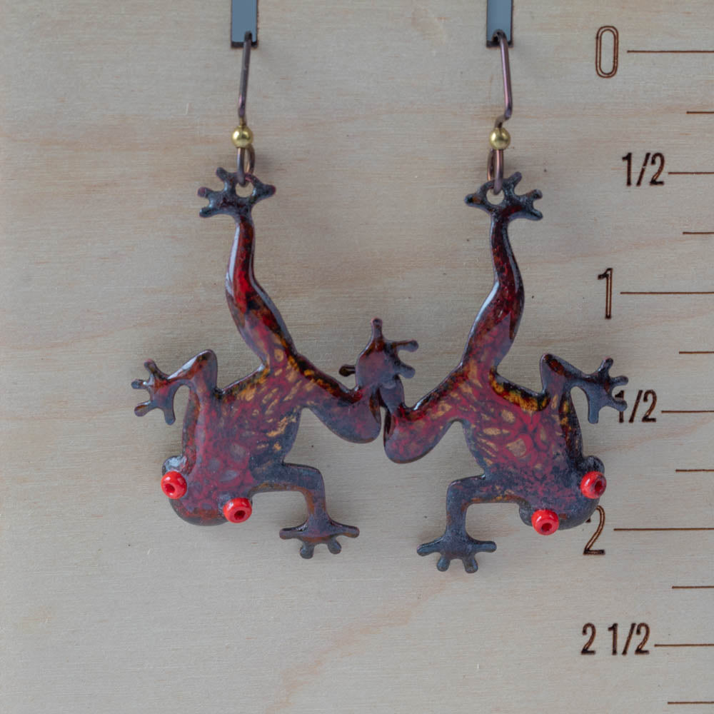 Metallic frog-shaped earrings on a wooden surface with measurement scale.