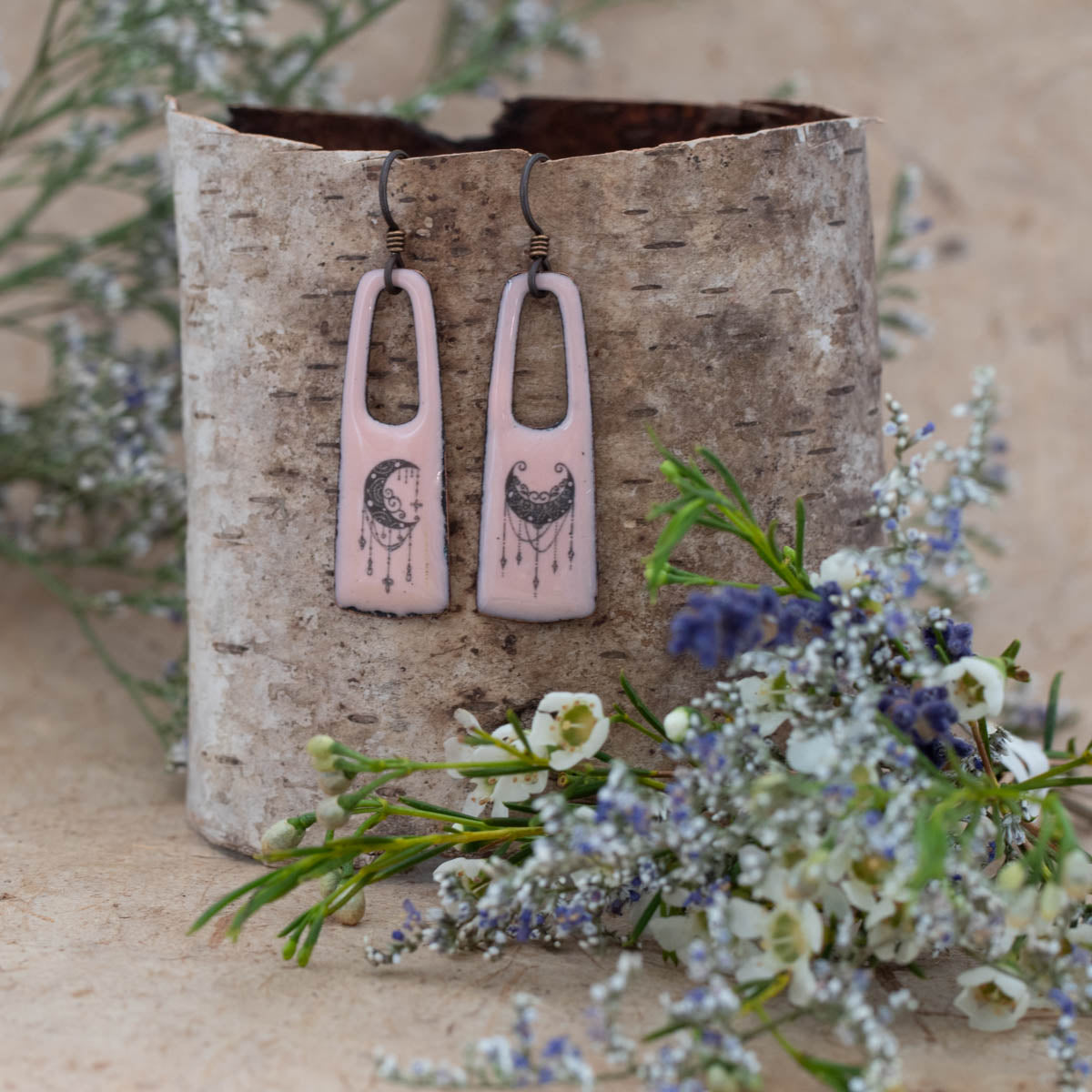 Romantic handmade enamel earrings in soft rose pink with black crescent moon and celestial designs, featuring long rectangular drop shapes with open tops. Displayed on birch bark with delicate wildflowers – Rose Moon Earrings by Divella Designs."