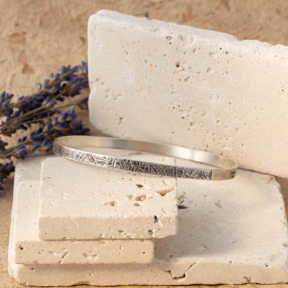Handmade sterling silver spiderweb textured bangle resting on stacked stone blocks with soft neutral tones and lavender in the background.