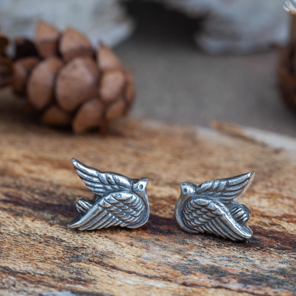 Handmade sterling silver dove stud earrings resting on rustic wood with pinecones in the background — nature inspired jewelry by Divella Designs.