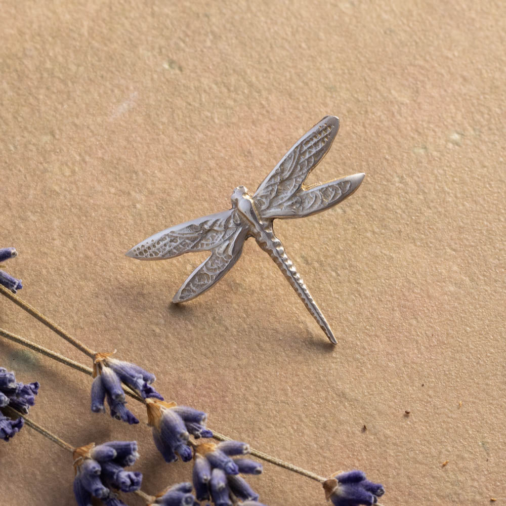 Silver dragonfly pin on a textured beige surface with lavender flowers.