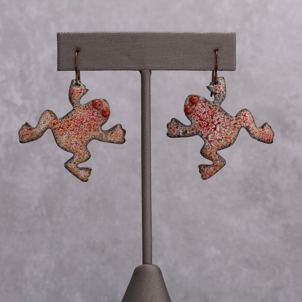 Playful red and green enamel frog-shaped earrings with dotted patterns and raised spiral eyes, displayed on a gray stand against a light gray background.