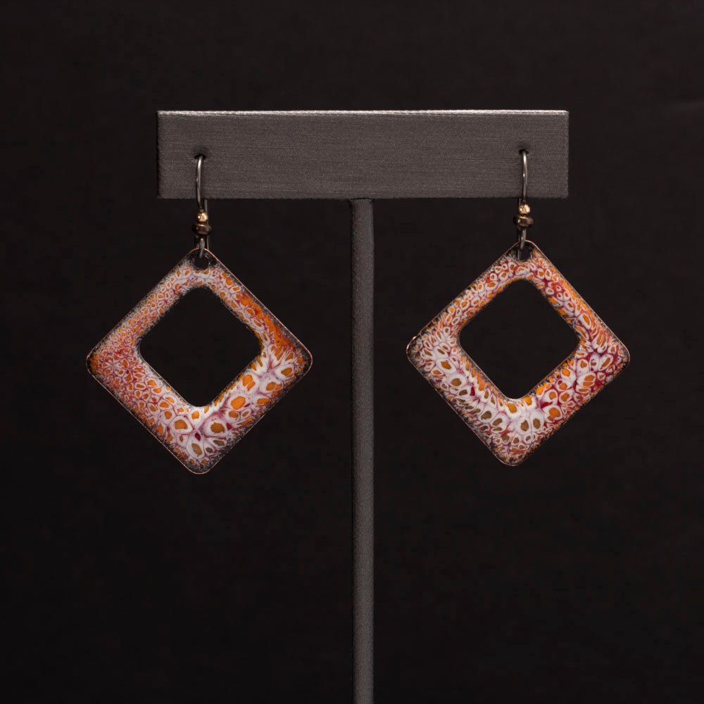 Square copper enamel earrings in earthy orange, rust, and white tones with an organic crackle pattern, displayed on an earrings display.
