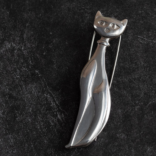 Handcrafted sterling silver cat brooch with a minimalist, elongated design and abstract facial features, displayed on a textured black background.