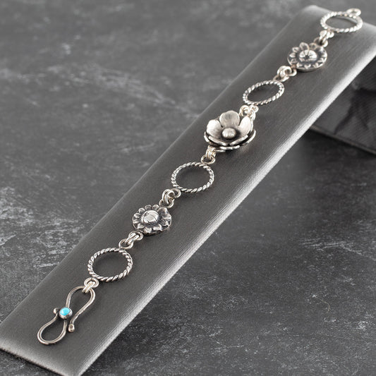 Handcrafted sterling silver bracelet featuring detailed floral charms, twisted rope links, and a turquoise-accented clasp – artisan botanical jewelry by Divella Designs, displayed on slate background.