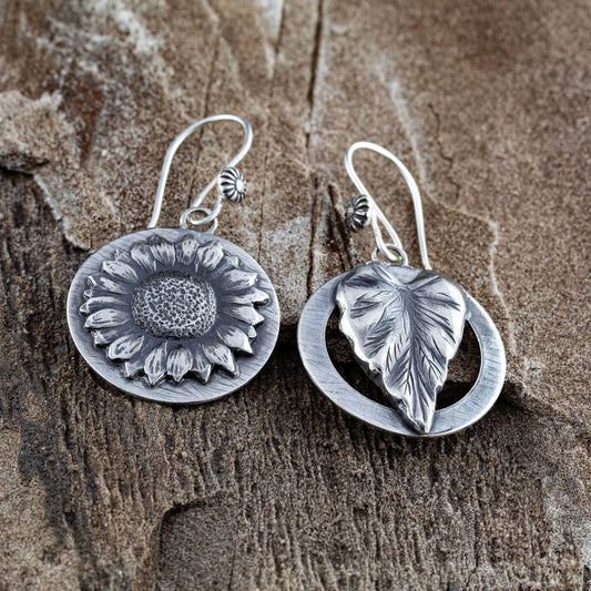 Sterling silver earrings with sunflower and leaf designs on round backings, featuring intricate hand-carved textures, displayed on rustic wood surface.