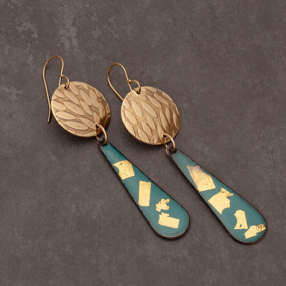 Teal enamel teardrop earrings adorned with 24k gold flakes and paired with round textured brass discs, displayed on a gray jewelry stand against a light gray background.