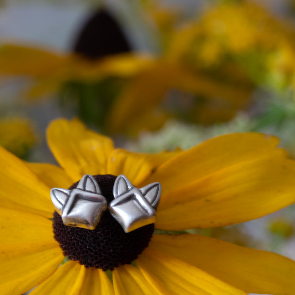 A pair of silver geometric stud earrings resting on the dark center of a yellow flower, with a softly blurred background of similar yellow flowers.