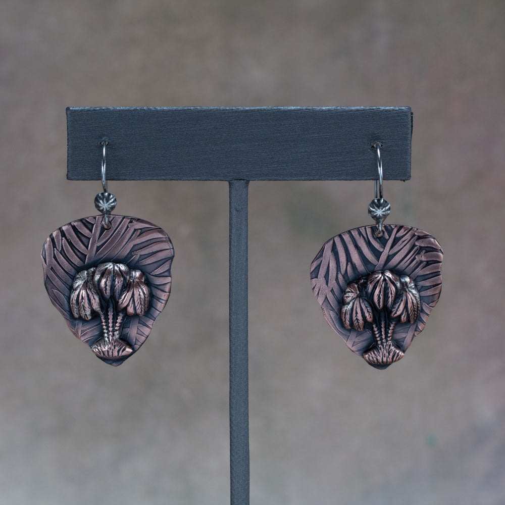 Handcrafted copper earrings with sculpted palm trees layered over embossed leaf-patterned backdrops, tropical artisan jewelry by Divella Designs – displayed on slate background