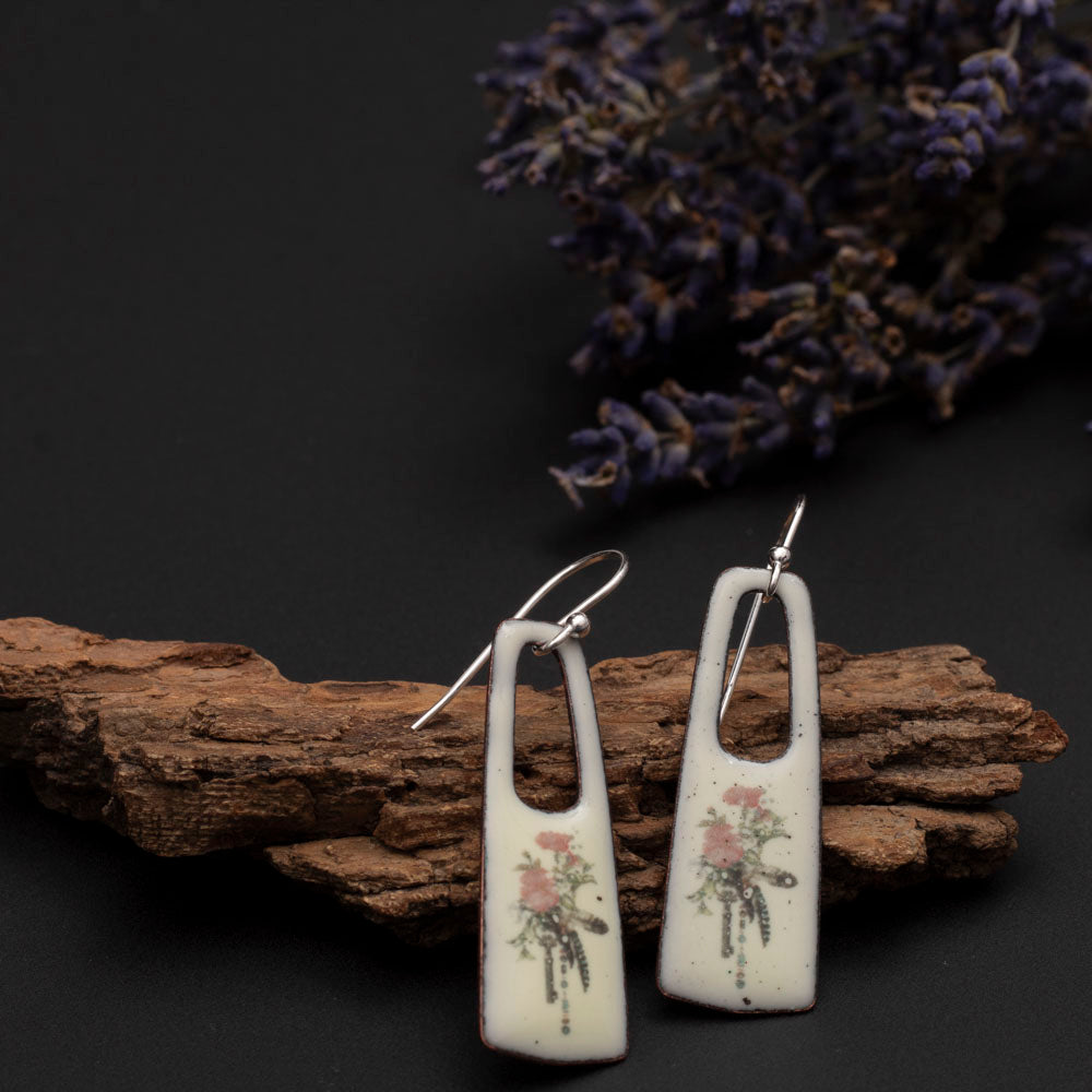Creamy enamel earrings with delicate pink floral bouquet designs, displayed on driftwood with lavender on a dark background.