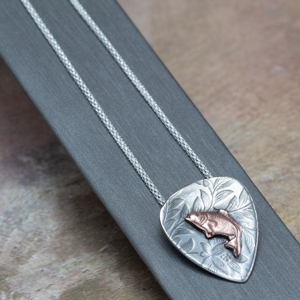 Handcrafted sterling silver pendant with etched floral background and raised copper jumping bass fish, hung on a silver chain – rustic nature-inspired necklace by Divella Designs, displayed on dark backgroun