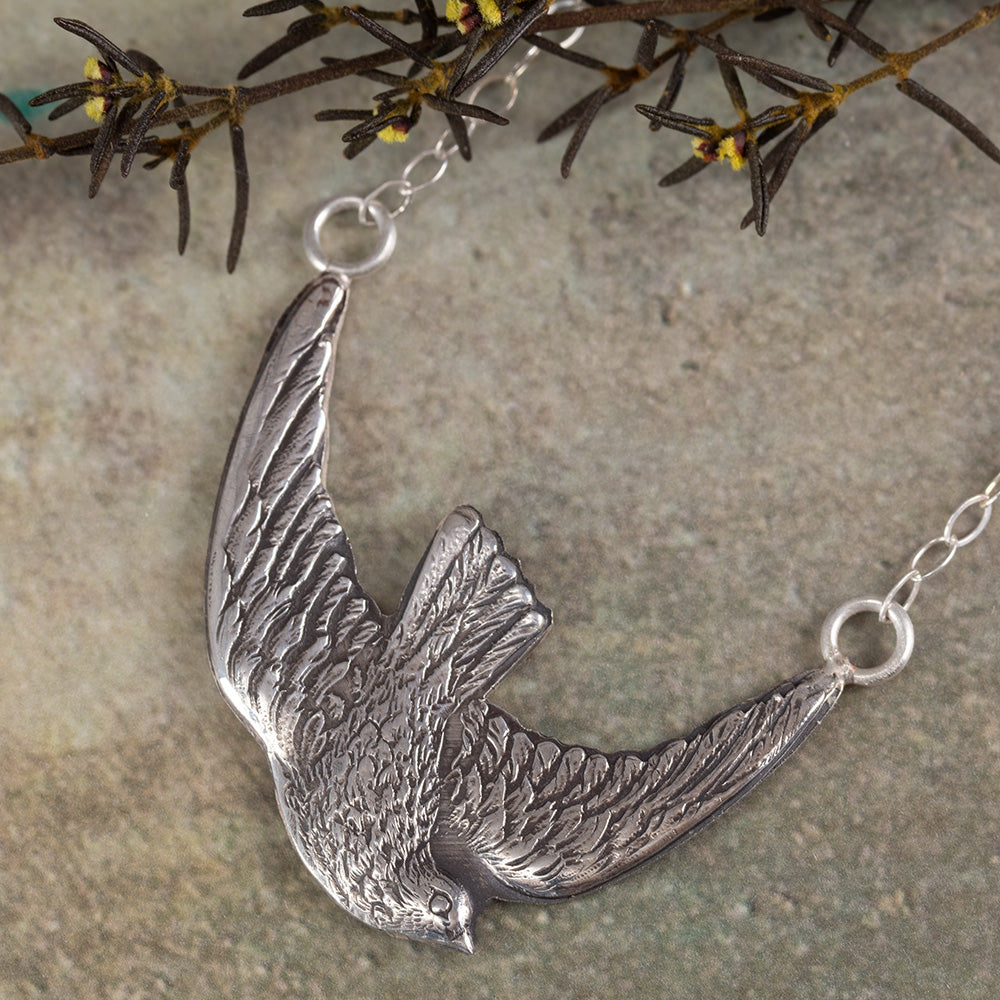 Sterling silver bird in flight pendant necklace with detailed feather texture, displayed on slate background with sprigs of greenery.