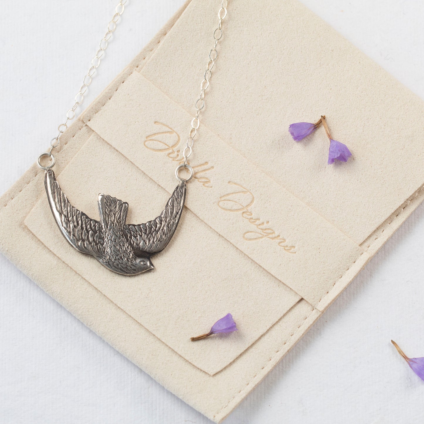 Sterling silver bird in flight pendant necklace with intricate feather detail, shown against a branded pouch with scattered purple flowers.