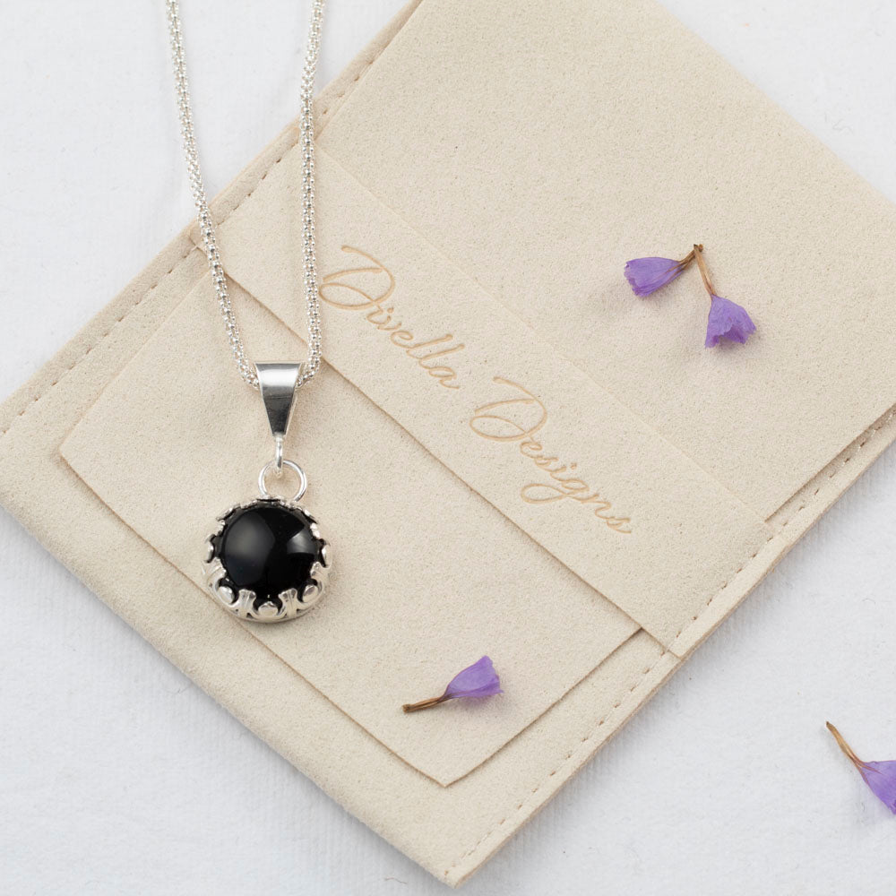 Sterling silver pendant necklace featuring a round black onyx stone in a scalloped bezel setting, displayed on a branded pouch with a white background with scattered purple flower petals.