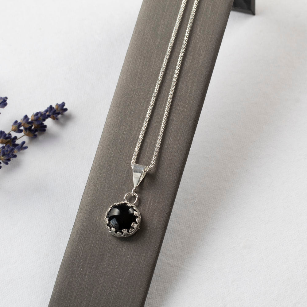 Sterling silver pendant necklace featuring a round black onyx stone in a scalloped bezel setting, displayed on a stand and white background with scattered purple flower petals.