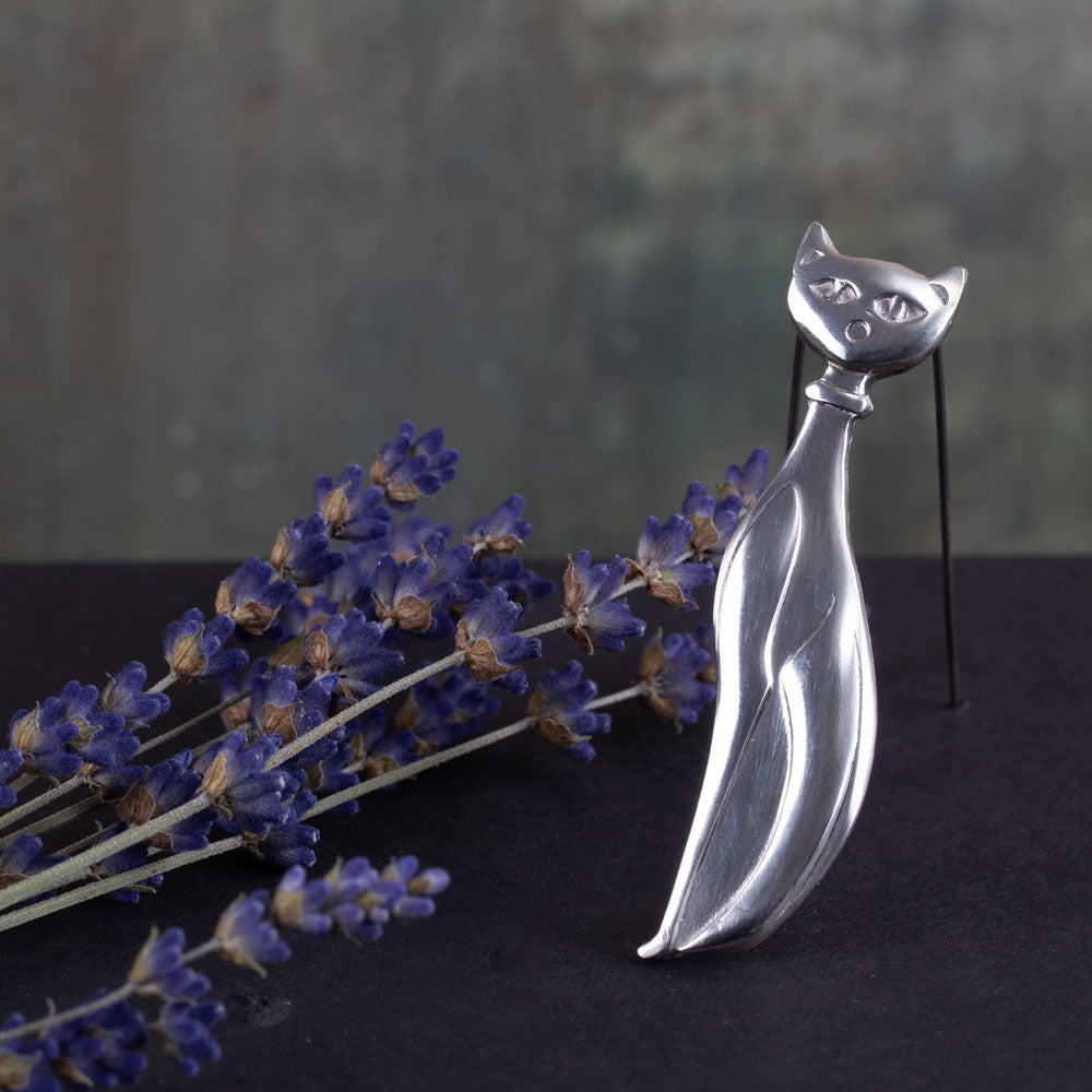 Elegant sterling silver cat brooch with a stylized, elongated body and minimalist features, displayed upright beside sprigs of dried lavender on a dark surface with a moody green-gray background.