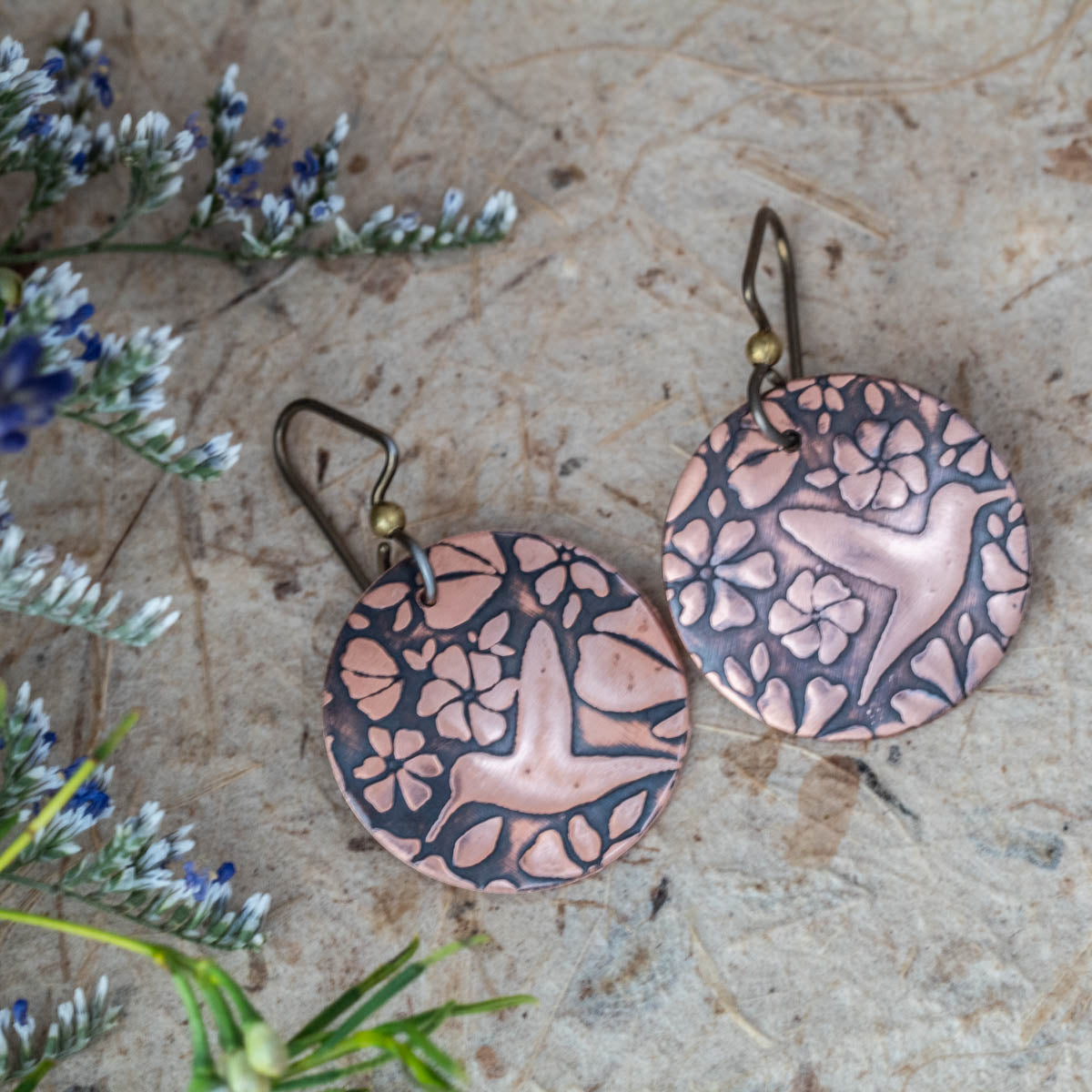 Handmade copper hummingbird earrings with floral motif, featuring etched round disc design and oxidized finish for a rustic, nature-inspired artisan look.
