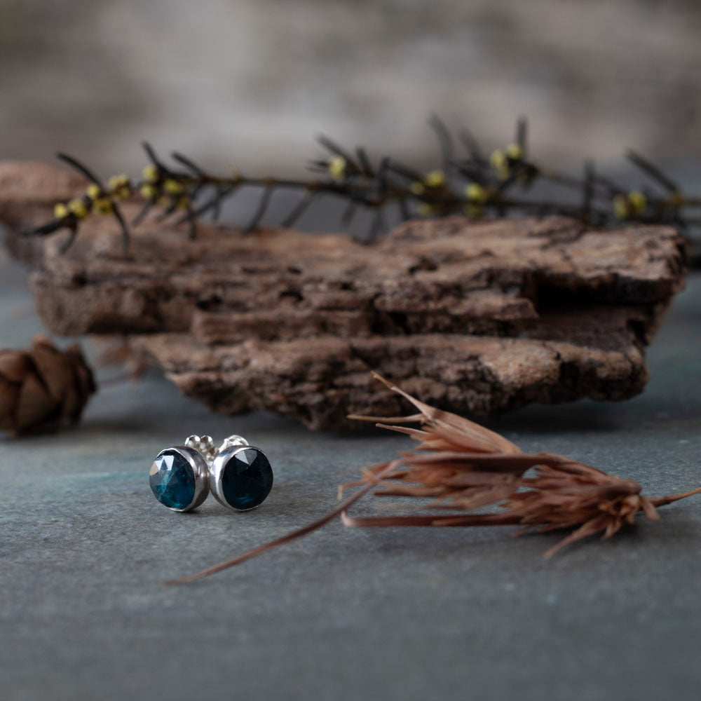Sterling silver stud earrings featuring faceted teal kyanite gemstones, photographed on piece of slate with bark and pine cones.