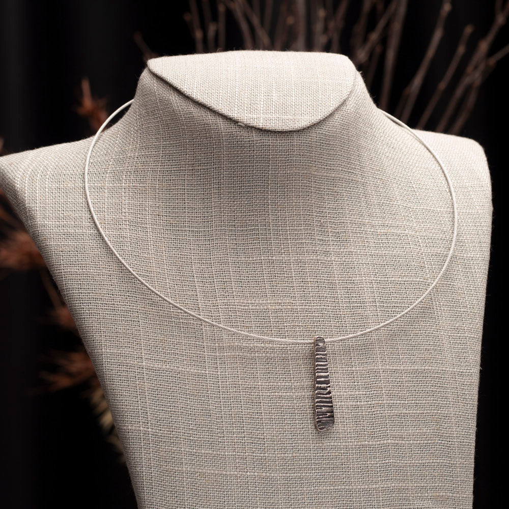 Minimalist silver pendant with tapered shape and organic linear texture on neck wire.