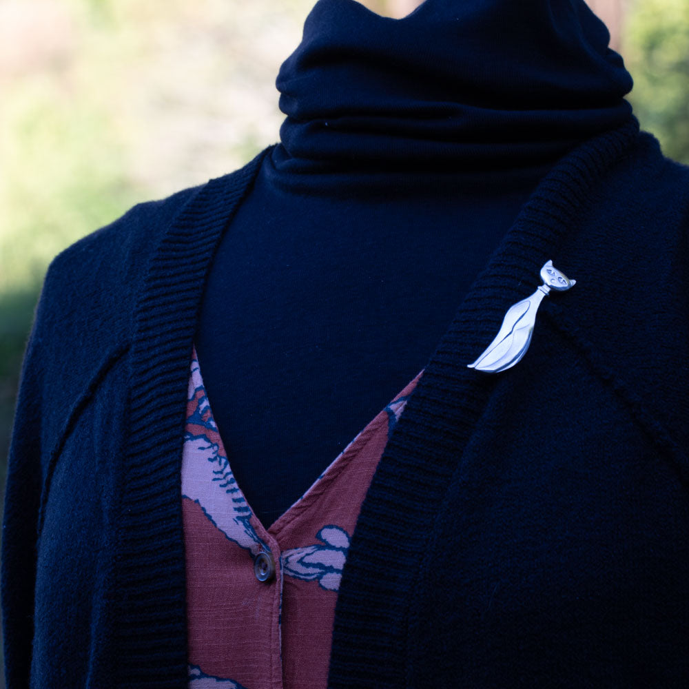Elegant sterling silver cat brooch with a stylized, elongated body and minimalist features, displayed on a mannequin wearing a black sweater and orange dress.