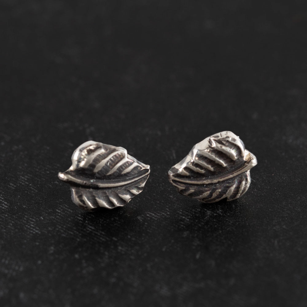 Tiny sterling silver leaf stud earrings with delicate vein detailing, nature-inspired minimalist design displayed on a black textured background – handmade by Divella Designs