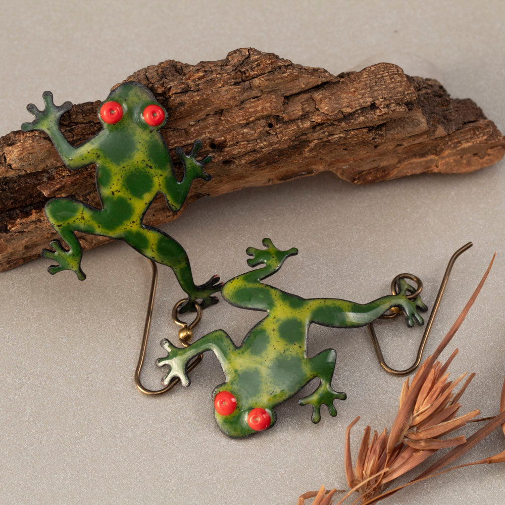 Playful green frog-shaped enamel earrings with bright red eyes, handcrafted and displayed on bark with woodsy bits – Enchanted Frog design by Divella Design