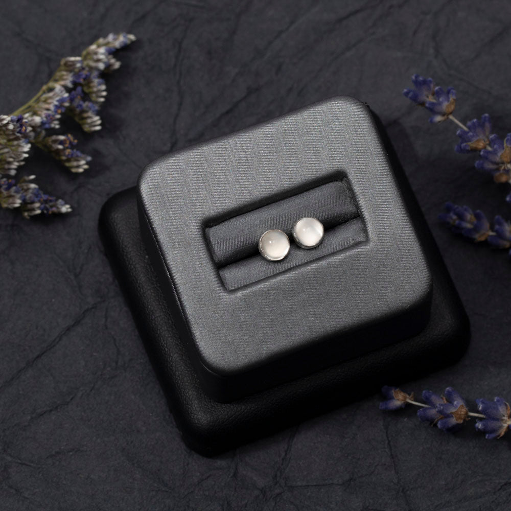 Round sterling silver moonstone stud earrings displayed on a black jewelry holder, surrounded by sprigs of lavender on a dark textured background