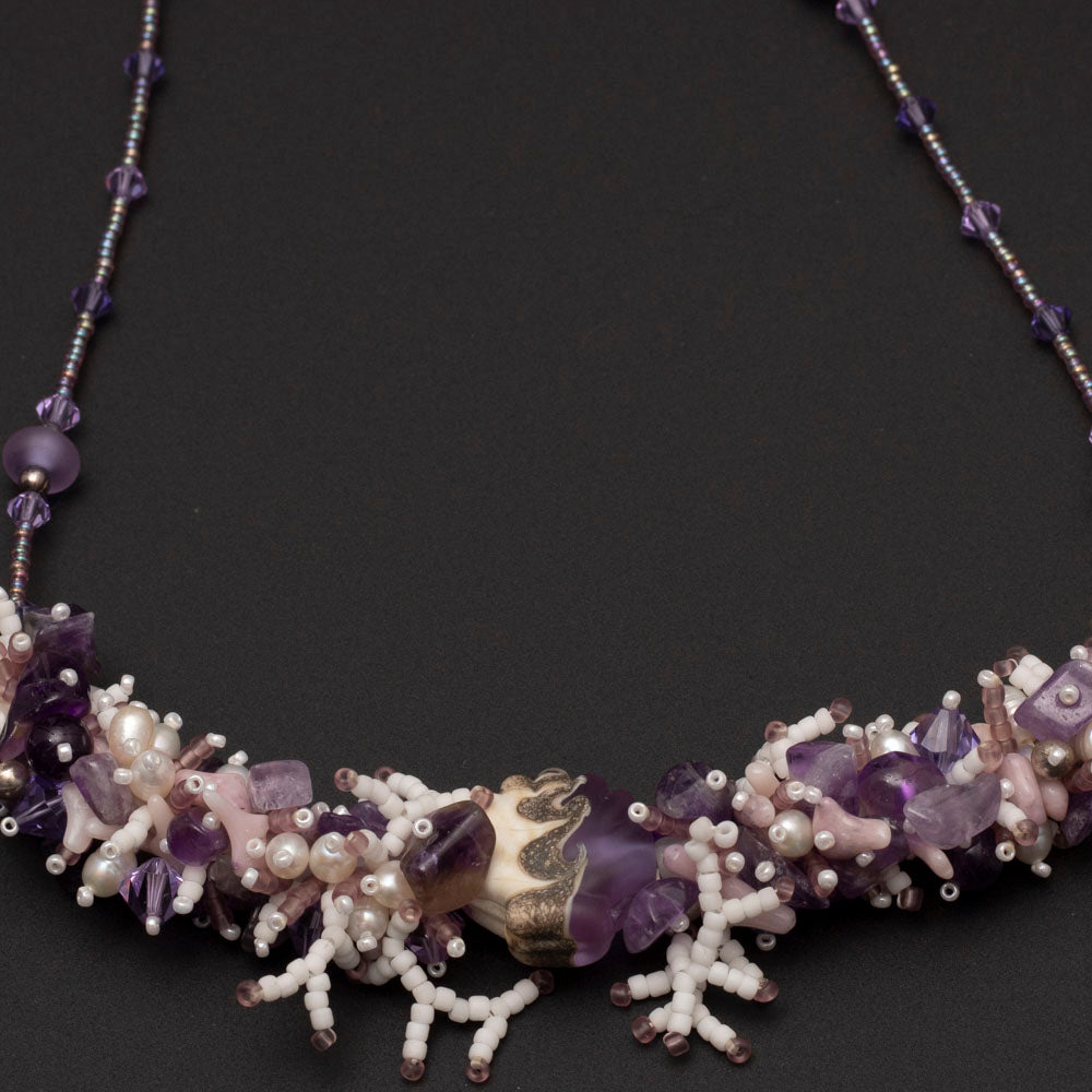 Close up of hand-beaded amethyst and pearl statement necklace with sculptural focal bead, styled on black background